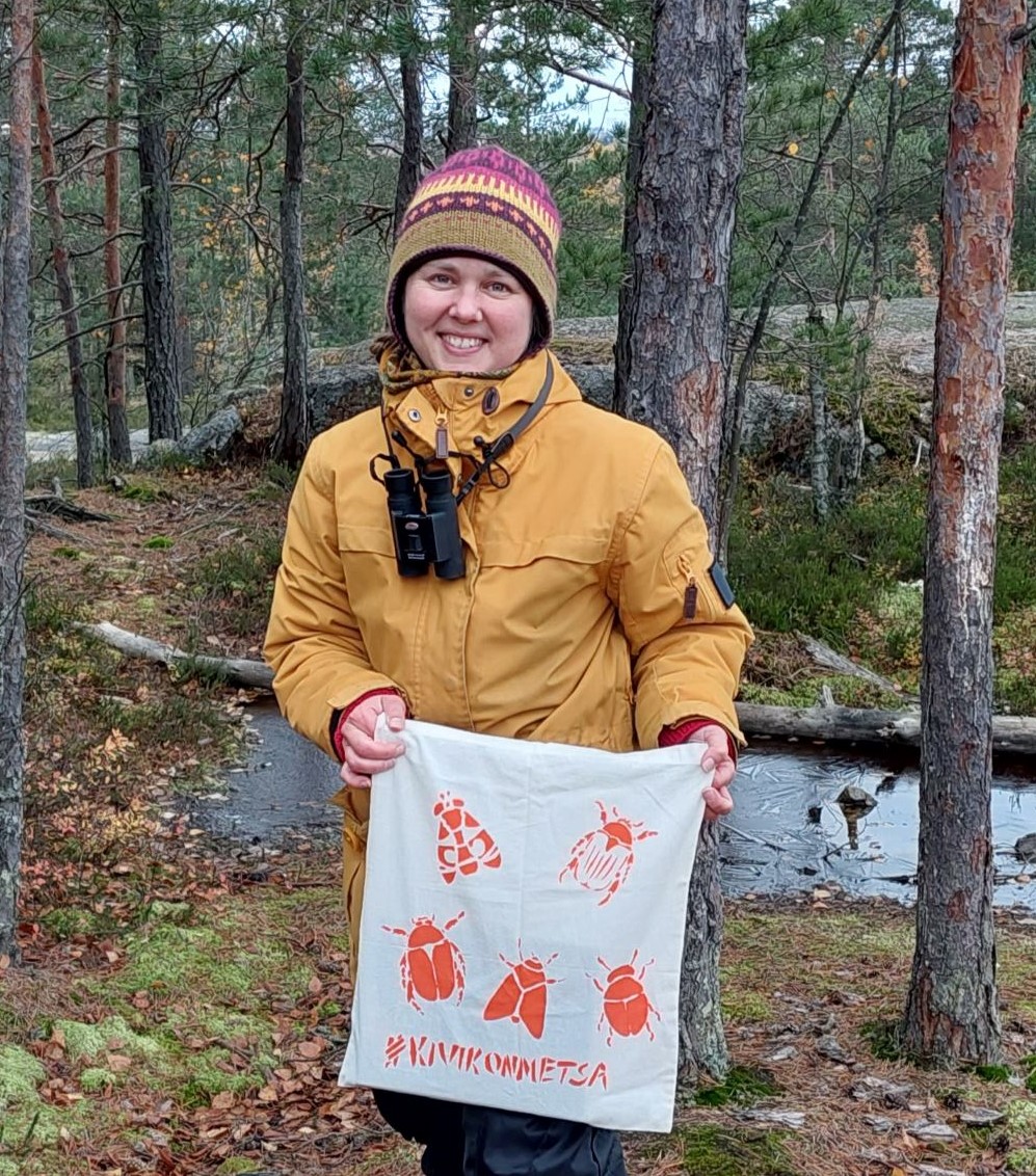 A woman in a forest, holding a fabric bag with text "Kivikon metsä", kivikko forest