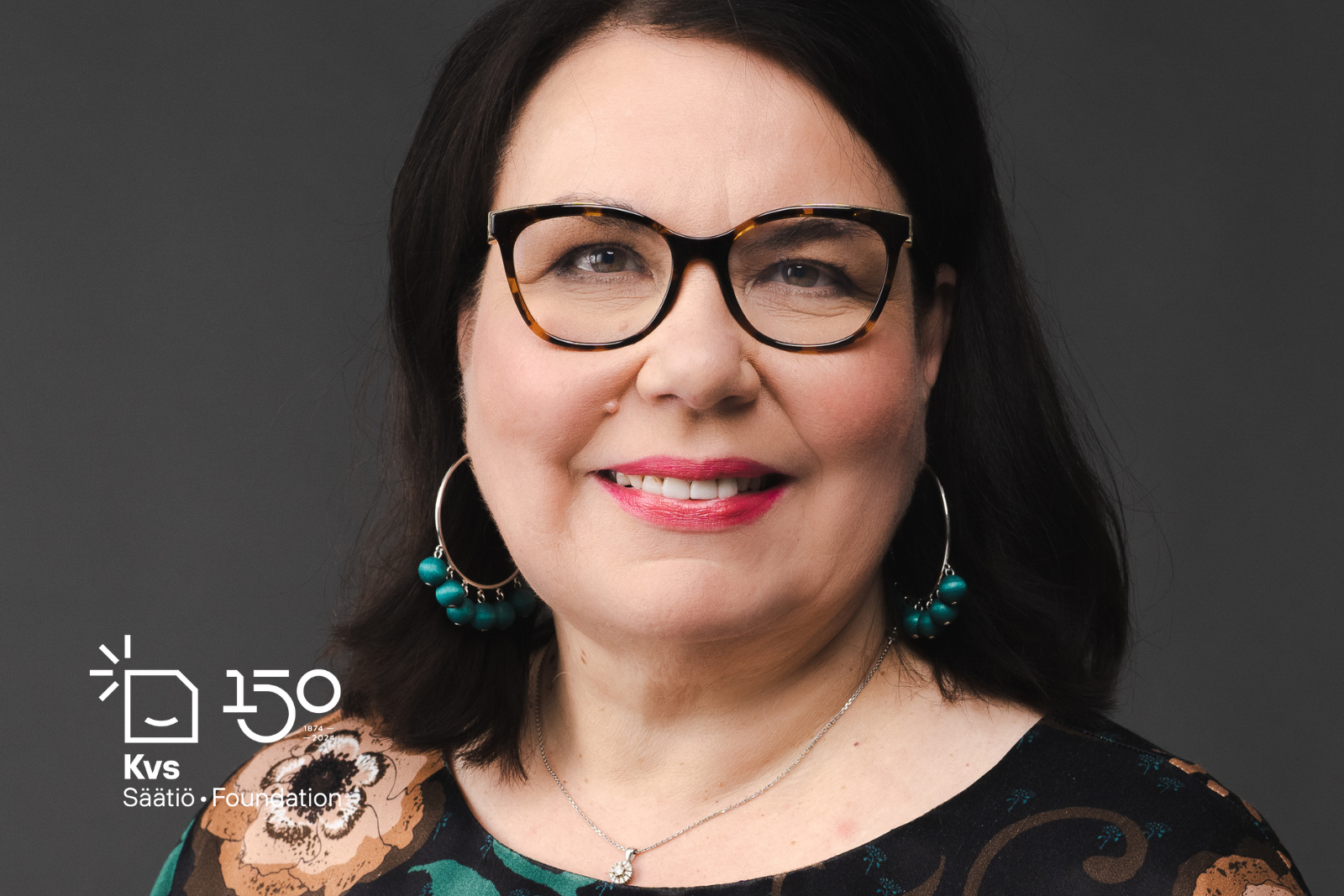 Kasvokuvassa hymyilevä tummatukkainen keski-ikäinen nainen tummasankaisissa silmälaisessa,