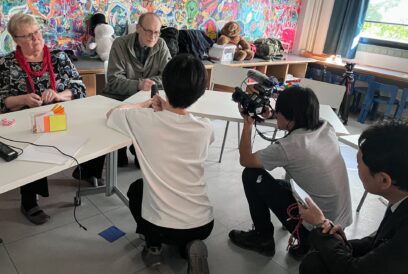 Valkoisen pöydän takana istuu ikääntynyt vaaleatukkainen nainen ja mies, jota kuvaa televisiokameralla kolme miestä.
