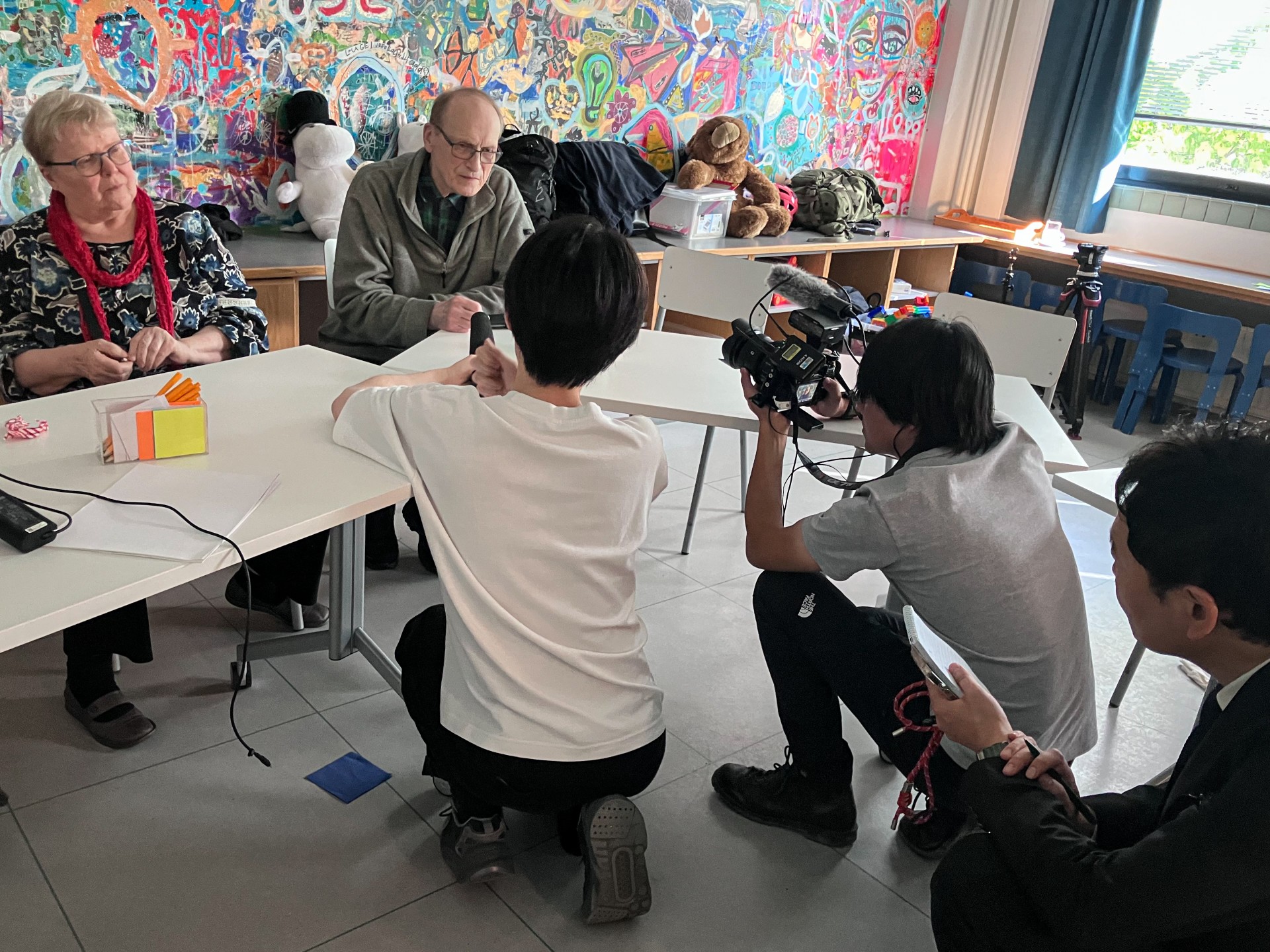 Valkoisen pöydän takana istuu ikääntynyt vaaleatukkainen nainen ja mies, jota kuvaa televisiokameralla kolme miestä.