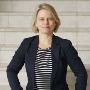 Vaaleatukkainen, tummaan bleiseriin ja raitapaitaan pukeutunut nainen kasvokuvassa.
