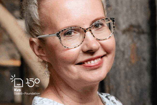 Vaaleatukkainen, muovisankaisiin silmälaseihin pukeutunut hymyilevä nainen tiukasti rajatussa kasvokuvassa.