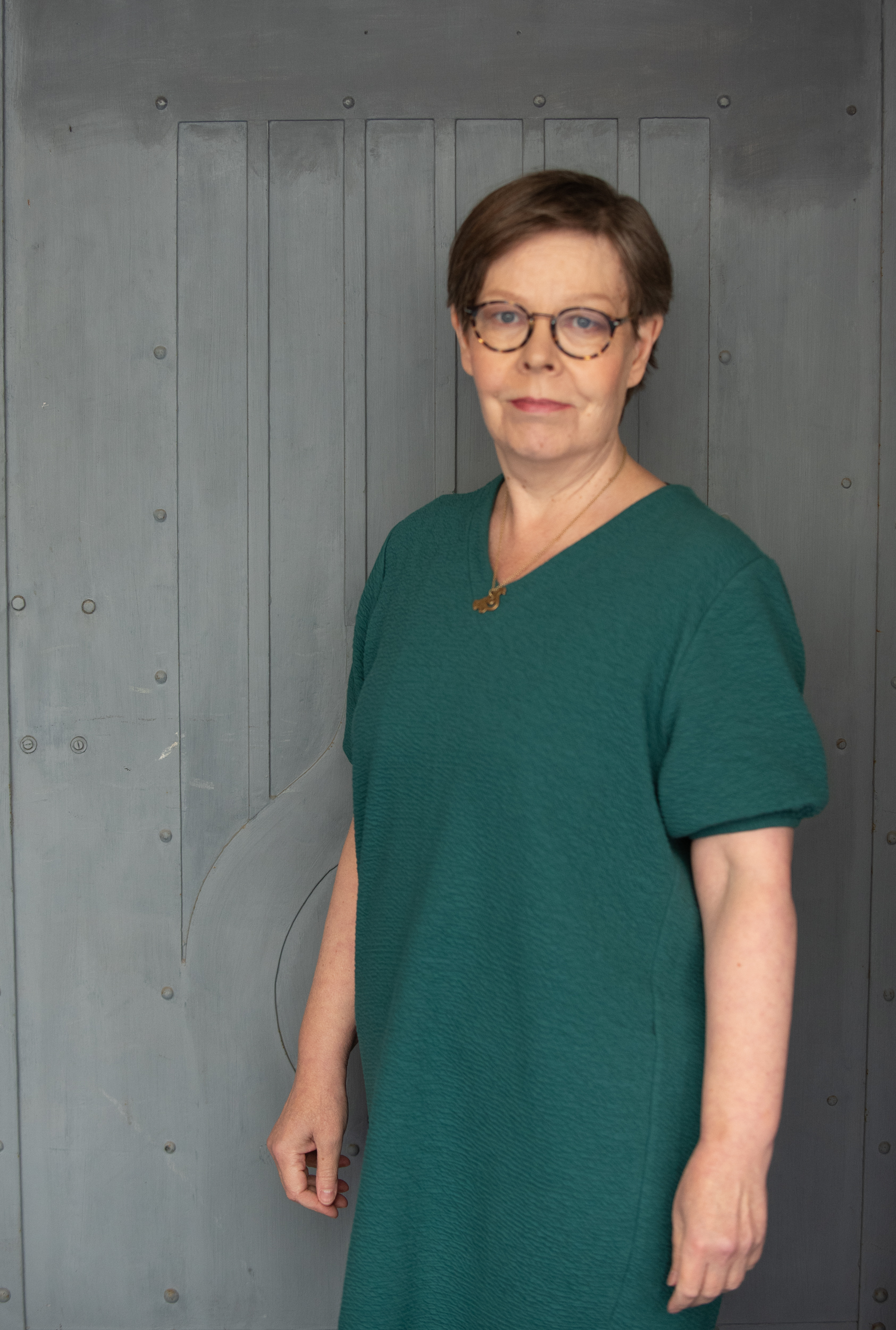 Vihreään asuun pukeutunut lyhyttukkainen silmälasipäinen nainen puolivartalokuvassa.