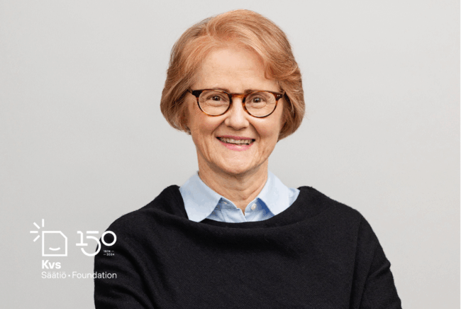 Hymyilevä punatukkainen, vaaleansiniseen paitapuseroon ja tummaan jumpperiin pukeutunut nainen kasvokuvassa.