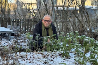 Silmälasipäinen lyhyttukkainen mies kyykyssä hennon lumen peittämässä puutarhassa.