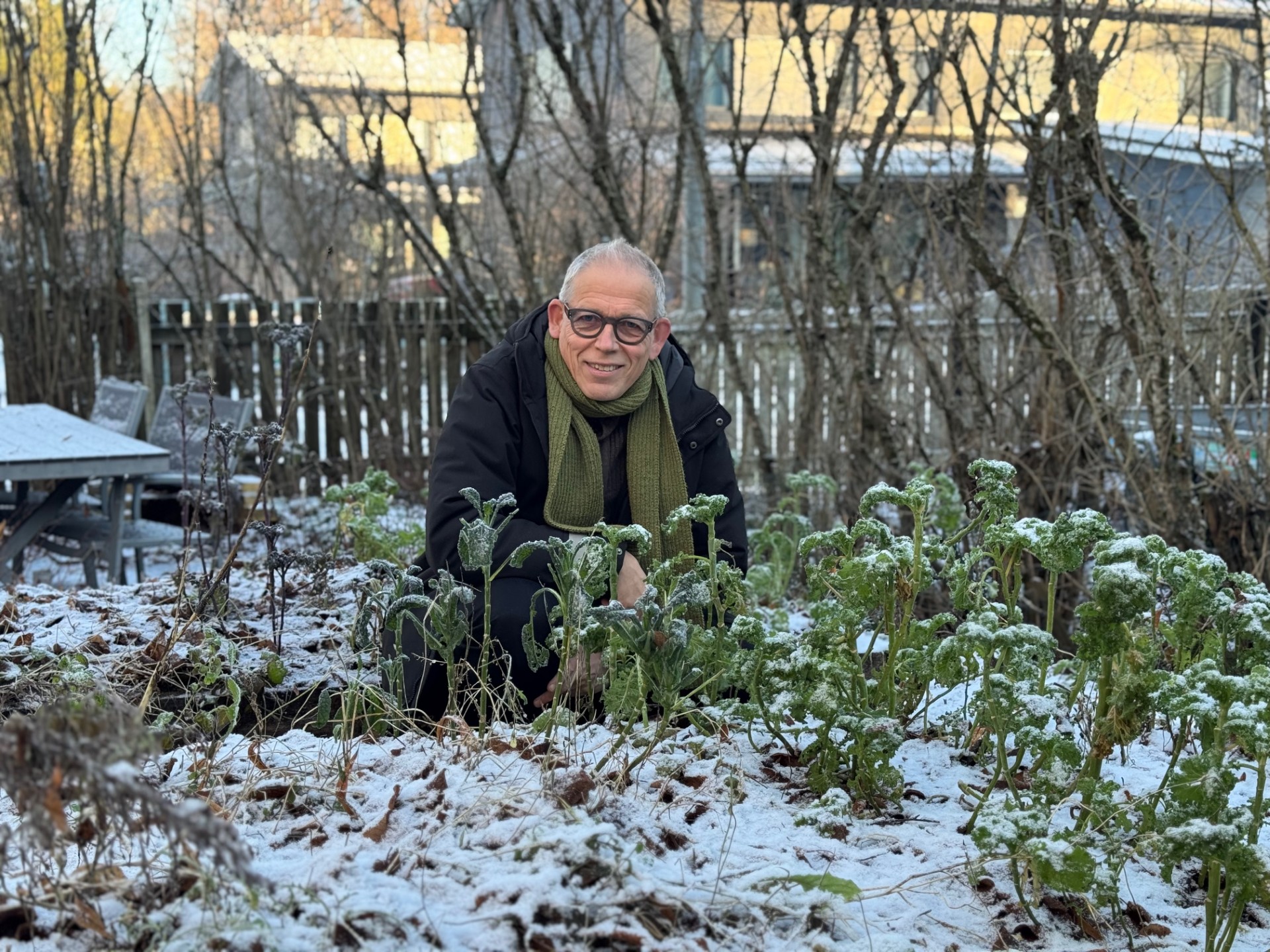Silmälasipäinen lyhyttukkainen mies kyykyssä hennon lumen peittämässä puutarhassa.